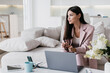 Serious pretty asian woman in business suit sitting on cozy couch at desk with laptop, phone, cup of coffee at home remote working. Attractive entrepreneur holding pen looking aside. Business people.