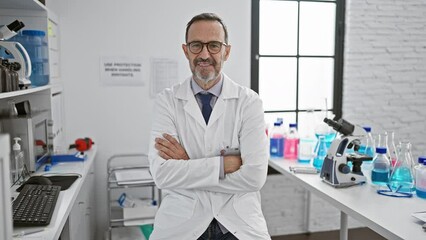 Poster - Happy mature, grey-haired male scientist with a confident smile enjoys his research work, sitting at his lab table, arms crossed in a welcoming gesture, connected with science and medicine.