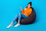 Fototapeta Panele - Photo of positive good mood woman dressed sweater sitting bean bag pointing two fingers empty space isolated blue color background