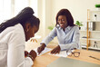 Portrait of smiling happy two business african american women working in office and signing a contract sitting at the desk on workplace making a deal and reaching agreement with colleague.