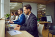 © Studio Romantic - Serious business people sitting in a row and working on laptop in modern open plan office sitting at same desk. Company employees wearing suits typing on computers on their workplace.