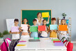 © Studio Romantic - Small group of school children together with teacher standing in classroom, holding different mockup posters and smiling. Happy junior students learn English and show colorful text copy space banners