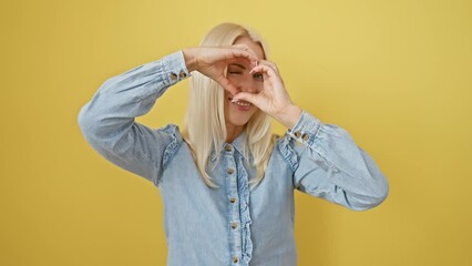 Canvas Print - Romantic concept, young blonde woman in denim shirt making love-heart symbol with hands, smiling cheerfully on isolated yellow background