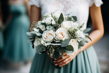 A bridal bouquet of white roses and greenery held by a bride with a bridesmaid in teal in the background.