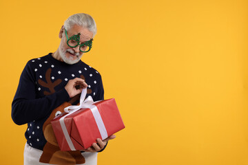 Canvas Print - Senior man in Christmas sweater and funny glasses opening gift against orange background. Space for text