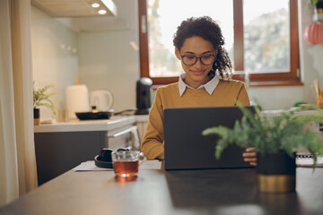 Smiling female manager working remotely on laptop from home while sitting near window