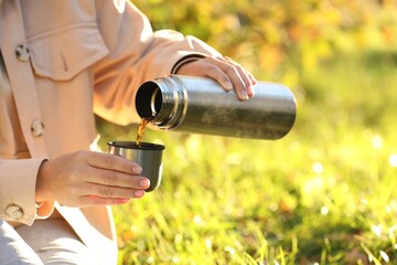 Wall Mural - Woman pouring tea from thermos into cup lid on green grass outdoors, closeup