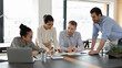 © fizkes - Happy multiethnic colleagues coworkers gather at desk in office discuss company financial paperwork together. Smiling multiracial businesspeople brainstorm collaborate with documents at workplace.