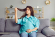 Portrait of a young brunette sad pregnant woman in casual clothes sitting on sofa in the living room at home and looking on her long hair falling out. Pregnancy and hair loss problems.