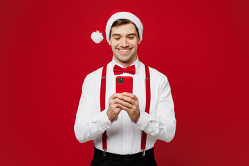 Wall Mural - Merry young cool fun man wears white shirt Santa hat posing hold in hand use mobile cell phone isolated on plain red background studio portrait. Happy New Year Christmas celebration holiday concept.