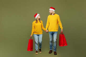 Full body merry cheerful young couple two friends man woman wears sweater Santa hat posing hold shopping bags isolated on plain light green background. Happy New Year Christmas sale buy day concept.