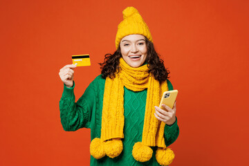 Young happy woman wear green knitted sweater yellow hat scarf using mobile cell phone hold credit bank card doing online shopping order delivery booking tour isolated on plain orange red background.