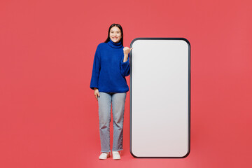 Wall Mural - Full body young woman of Asian ethnicity wear blue sweater casual clothes point thumb finger on big huge blank screen mobile cell phone smartphone with area isolated on plain pastel pink background.