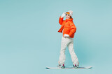 Full body sideways young woman wear warm padded windbreaker jacket hat ski goggles mask snowboarding look camera travel rest spend weekend winter season in mountains isolated on plain blue background