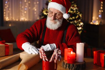 Poster - Santa Claus wrapping gift at his workplace in room decorated for Christmas