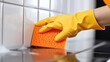 Close-up of a hand in a yellow cleaning glove holding an orange sponge, wiping down white ceramic tiles.
