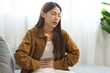 © KMPZZZ - Flatulence asian young woman hand in stomach ache, suffer from food poisoning, abdominal pain and colon problem, gastritis or diarrhoea sitting on sofa at home. Patient belly, abdomen or inflammation.