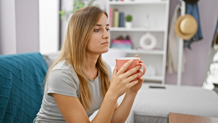 Canvas Print - Attractive young blonde woman comfortably sitting indoors in her apartment's living room, enriching her morning by relaxing and drinking a warm espresso coffee while doubtingly pondering.