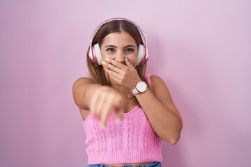 Sticker - Young blonde woman listening to music using headphones laughing at you, pointing finger to the camera with hand over mouth, shame expression