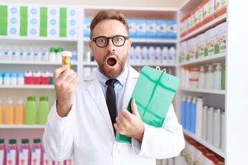 Poster - Middle age man with beard working at pharmacy drugstore holding pills in shock face, looking skeptical and sarcastic, surprised with open mouth