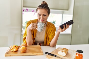 Poster - Young beautiful hispanic woman having breakfast drinking coffee using smartphone at the kitchen