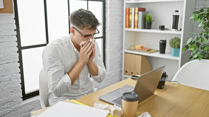 Young, handsome caucasian man, a focused business worker, battling sickness while crunching data on his laptop at the office, despite a relentless flu.