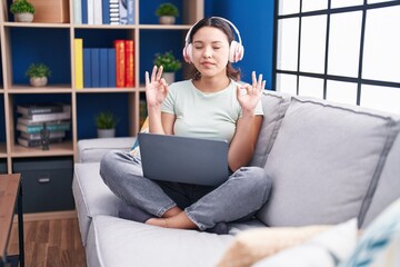 Sticker - Hispanic young woman using laptop at home wearing headphones relax and smiling with eyes closed doing meditation gesture with fingers. yoga concept.