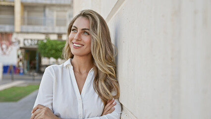 Sticker - Beautiful young blonde woman happily standing at city street corner, confidently looking to the side with arms crossed, emanating positivity and joy while warmly smiling outdoors.