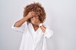 Young hispanic woman with curly hair standing over white background covering eyes and mouth with hands, surprised and shocked. hiding emotion