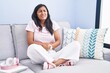 © Krakenimages.com - Young hispanic woman sitting on the sofa at home with hand on stomach because nausea, painful disease feeling unwell. ache concept.