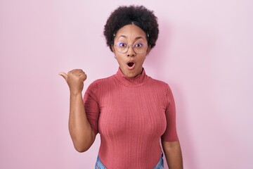 Sticker - Beautiful african woman with curly hair standing over pink background surprised pointing with hand finger to the side, open mouth amazed expression.