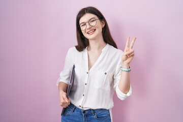 Wall Mural - Young caucasian woman holding laptop smiling looking to the camera showing fingers doing victory sign. number two.