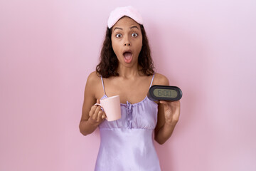 Wall Mural - Young hispanic woman wearing nightgown holding alarm clock afraid and shocked with surprise and amazed expression, fear and excited face.