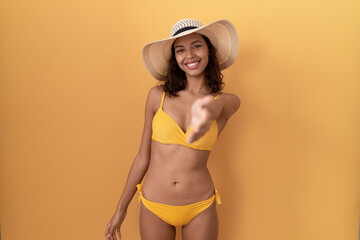 Wall Mural - Young hispanic woman wearing bikini and summer hat smiling friendly offering handshake as greeting and welcoming. successful business.