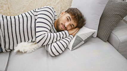 Poster - Cheerful, relaxed young arab man lying confidently on a cozy sofa, smiling joyfully at home. relishing the comfort of relaxation indoors, showcasing a happy, positive expression.