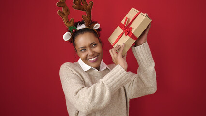 Poster - African american woman listening sound christmas gift wearing reindeer ears over isolated red background