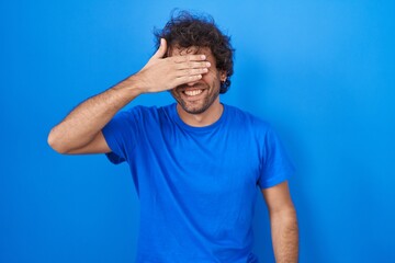 Sticker - Hispanic young man standing over blue background smiling and laughing with hand on face covering eyes for surprise. blind concept.