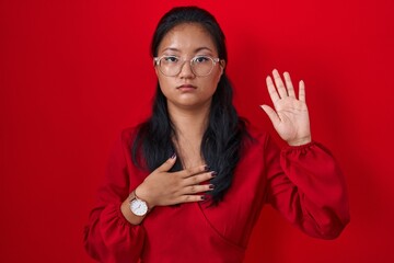 Wall Mural - Asian young woman standing over red background swearing with hand on chest and open palm, making a loyalty promise oath