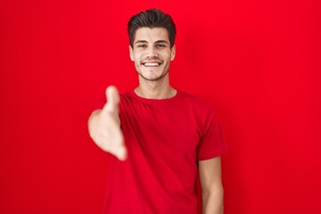 Poster - Young hispanic man standing over red background smiling friendly offering handshake as greeting and welcoming. successful business.