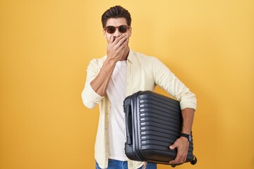 Canvas Print - Young hispanic man holding suitcase going on summer vacation laughing and embarrassed giggle covering mouth with hands, gossip and scandal concept