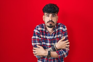 Wall Mural - Young hispanic man with beard standing over red background shaking and freezing for winter cold with sad and shock expression on face