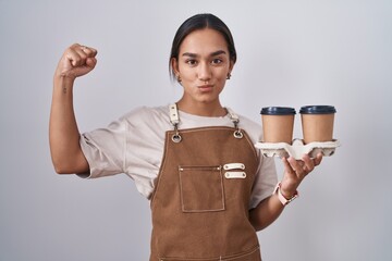 Wall Mural - Young hispanic woman wearing professional waitress apron holding coffee strong person showing arm muscle, confident and proud of power