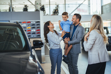 Wall Mural - Young happy family enjoying while buying a new car in a showroom. Saleswoman at car dealership center helping family to choose new family vehicle. Family in a car dealership