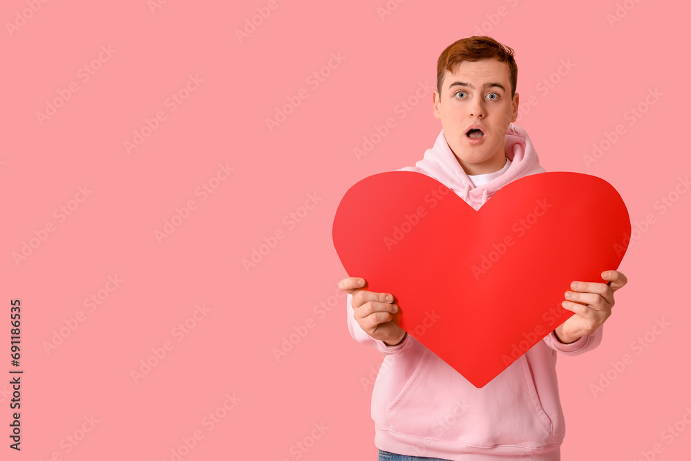Shocked young man with red paper heart on pink background