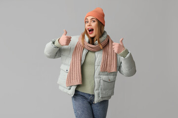 Poster - Beautiful young shocked woman in warm winter clothes showing thumbs-up gesture on grey background