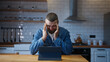 Bearded young adult man sitting against the kitchen counter having headache while using tablet device. Tired young man get having pain from a headache or migraine