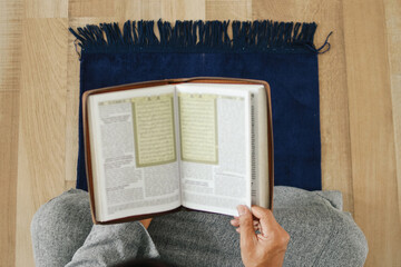 Wall Mural - Top view and close up of muslim man holding holy book of Quran or Koran while sitting on prayer mat