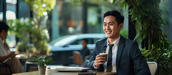 Poster - A young Asian man in business attire sits at an outdoor cafe, smiling and talking on his phone while enjoying a cup of coffee.