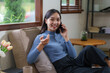 © amnaj - Attractive Asian woman sitting on sofa talking on phone at home.