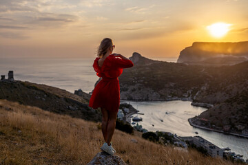 Wall Mural - Happy woman standing with her back on the sunset in nature in summer with open hands posing with mountains on sunset, silhouette. Woman in the mountains red dress, eco friendly, summer landscape